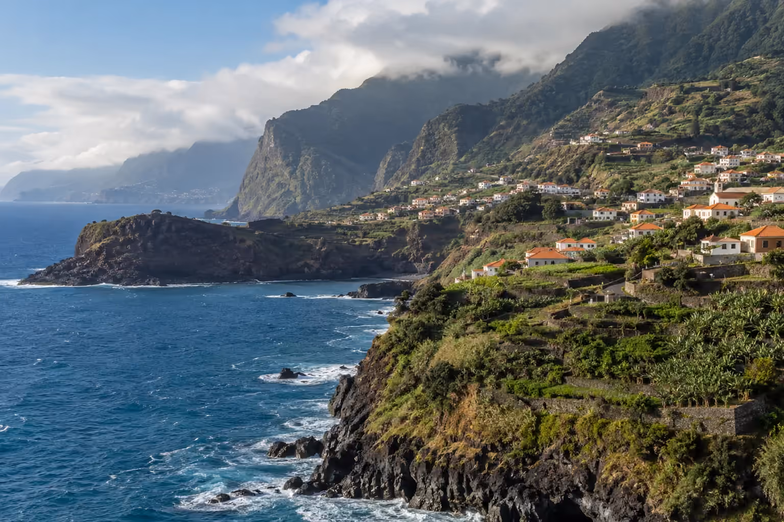 Green terraced cliffs and white houses above blue Atlantic water, with low clouds over the mountains