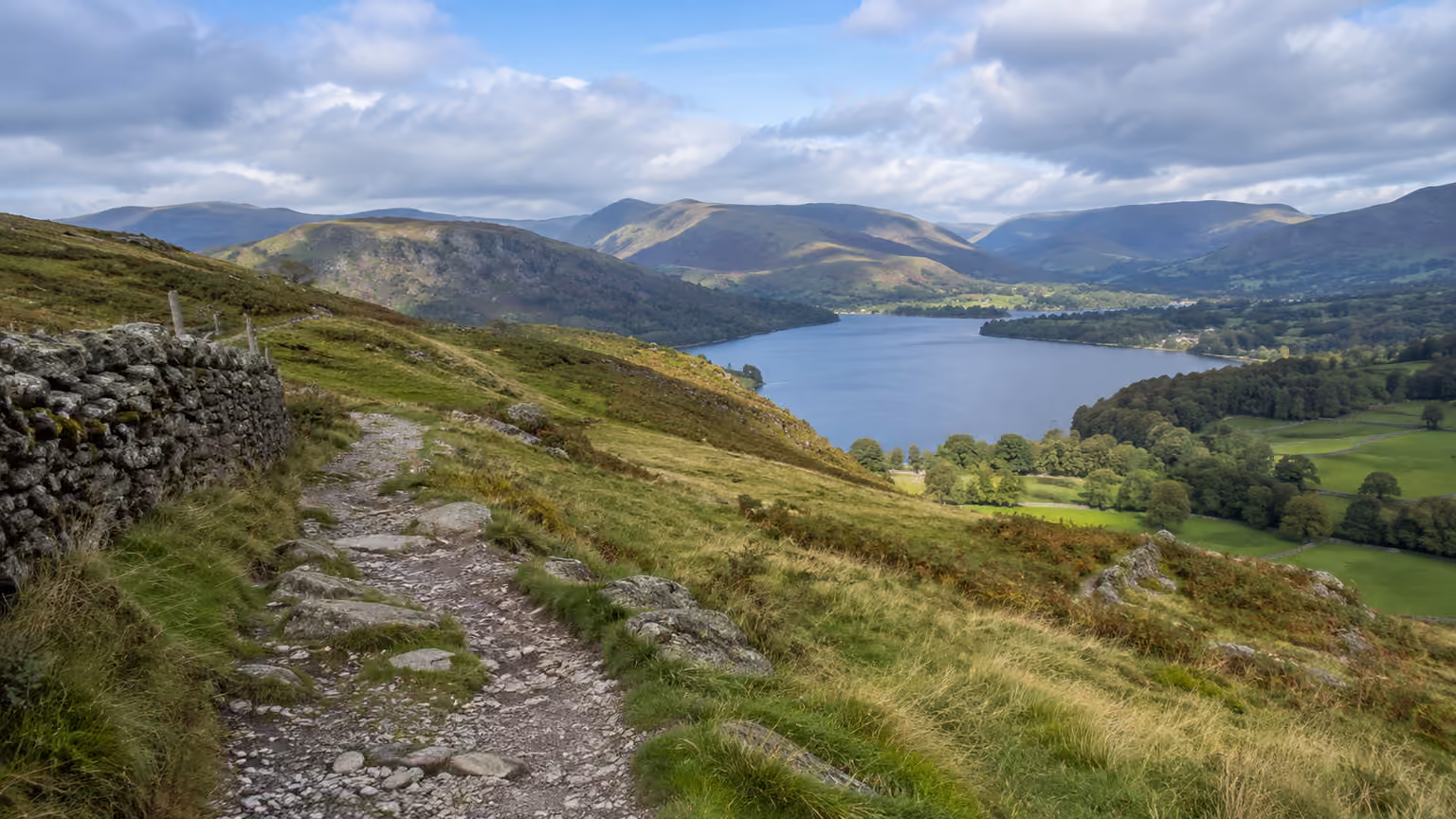 A public footpath through grassy Lake District fells with a distant lake and weathered stone walls