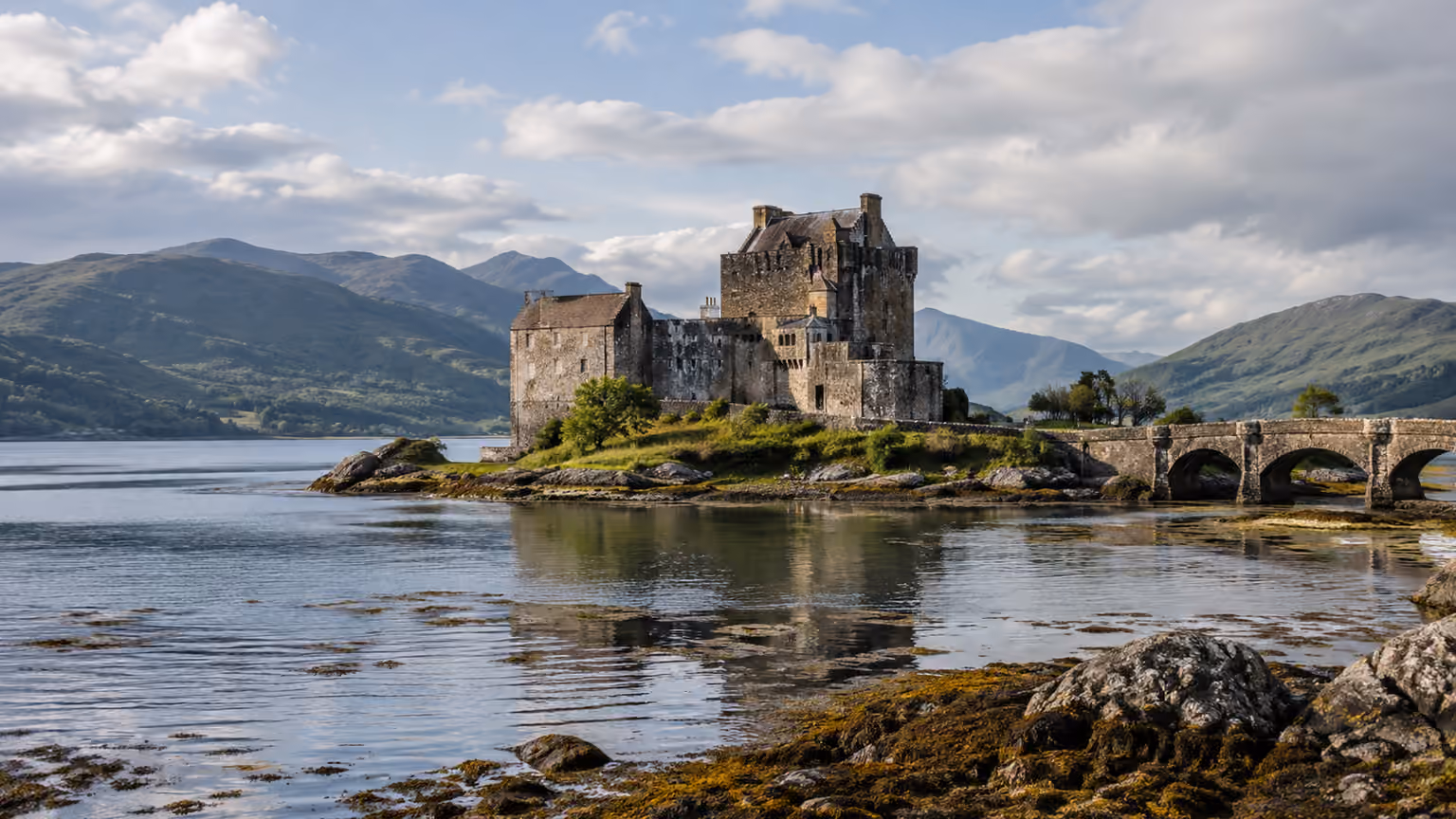A stone castle and arched bridge on a tidal island beneath green Highland hills