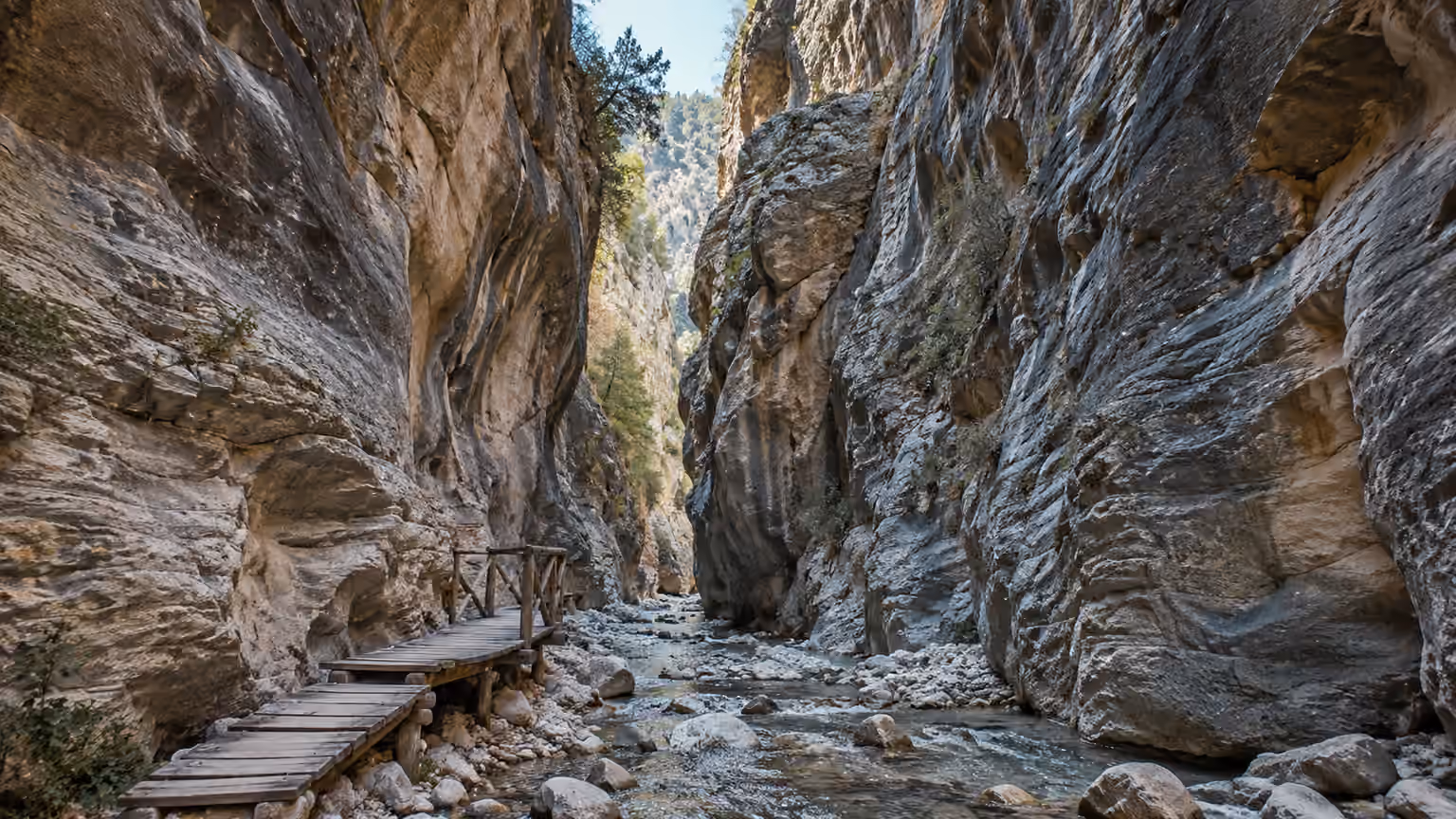 Tall weathered limestone canyon walls beside a rocky stream bed and wooden footbridge